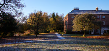 Tapton house winter This landscape photograph captures Tapton House during a clear winter morning, with the grounds of Tapton Park in Chesterfield, United Kingdom visible in the foreground. Nature dominates the scene, with bare trees and scattered leaf litter across the frosty grass, highlighting the seasonal change. Tapton House, the main subject of the image, is a well-known building and landmark within the park, and stands prominently to the right, partially framed by mature trees. The early sunlight emphasizes the frost and shadows on the ground, enhancing the wintry atmosphere of the park. The presence of benches along the park path suggests a public space designed for visitors to enjoy the natural setting and historic architecture. The image represents a typical winter landscape found in British parks such as Tapton Park, blending trees, nature, and local history.